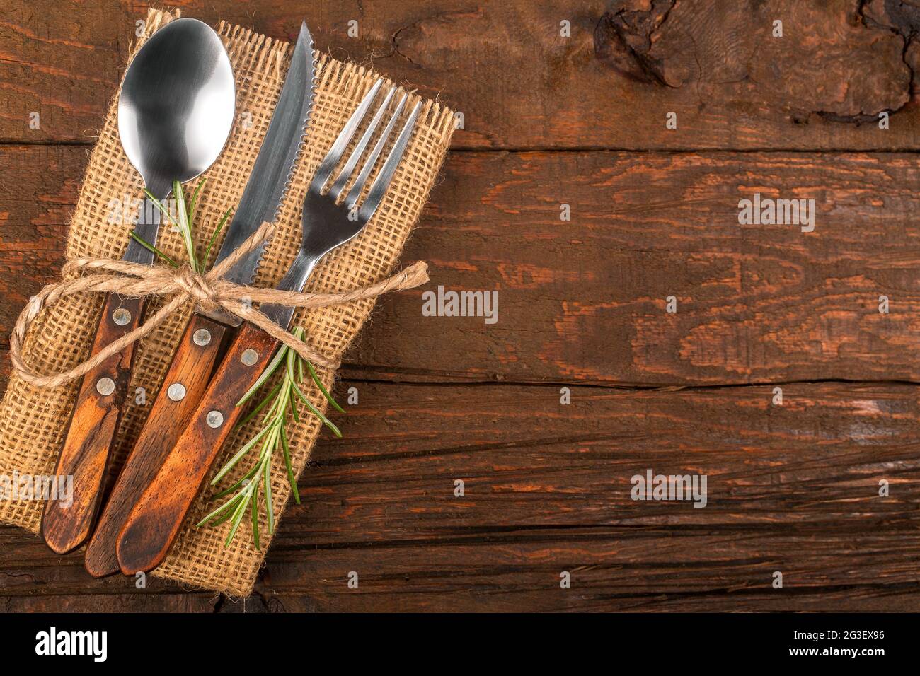 Posate con tovagliolo di lino e decorazione di rosmarino fresco su una base rustica in legno. Un cucchiaio, forchetta e coltello come utensili da cucina e da cucina. Foto Stock