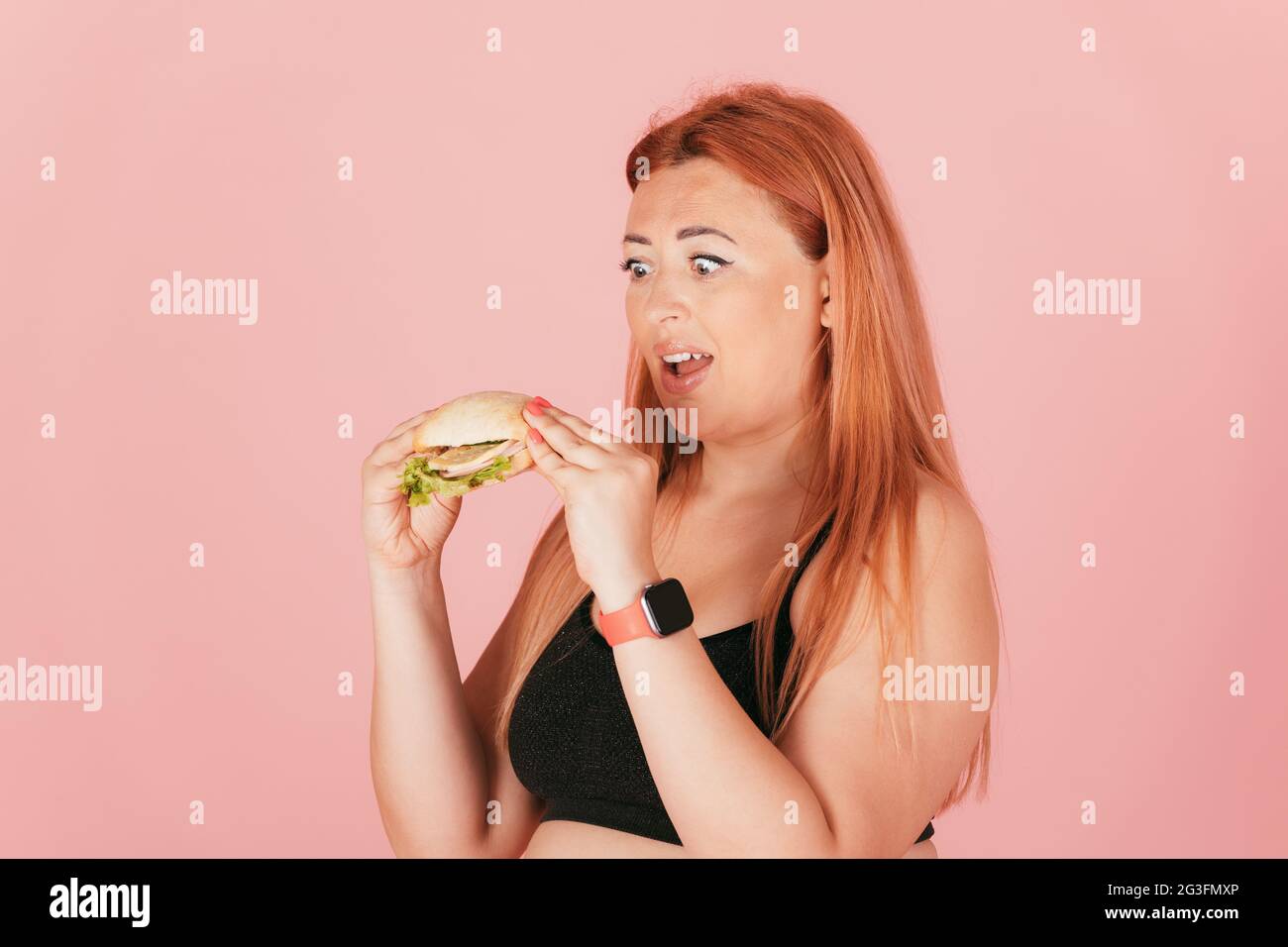 Affamato rosso donna sovradimensionata mangiare fast food hamburger su sfondo rosa. Foto Stock