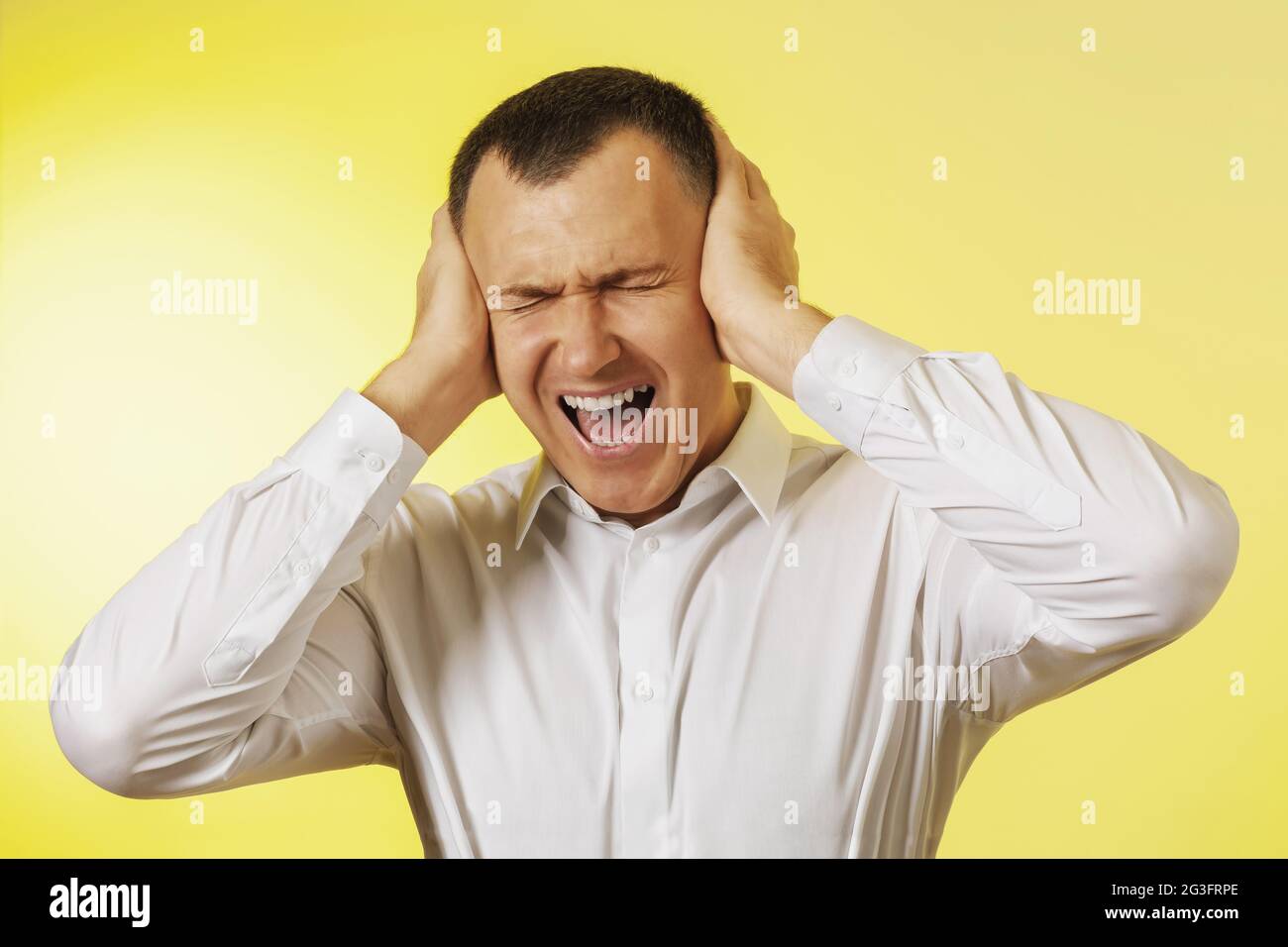 Un uomo grida coprendo le orecchie con le mani su uno sfondo giallo, il concetto di mal di testa Foto Stock