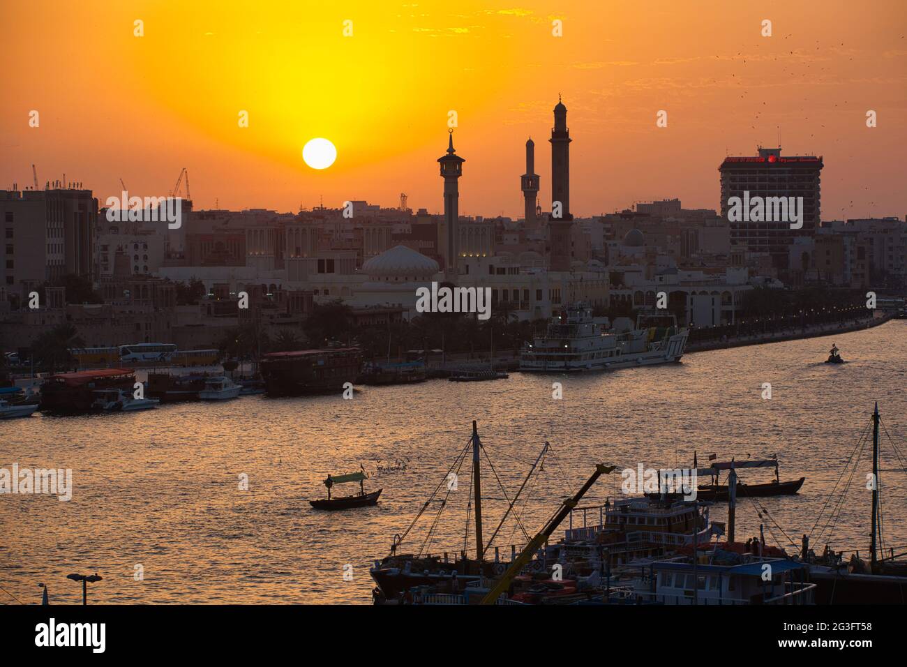 Uno skyline della città di Dubai al tramonto con tre minareti in silhouette e un grande edificio contro il cielo arancione. Il sole che tramonta, Dubai Creek. Foto Stock