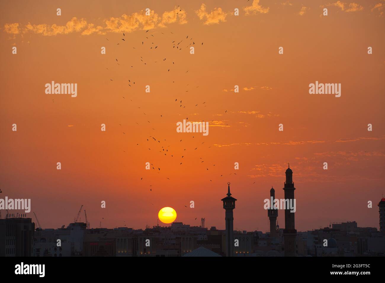 Uno skyline della città di Dubai al tramonto con tre minareti in silhouette e un grande gregge di uccelli contro il cielo arancione. Il sole che scende Foto Stock