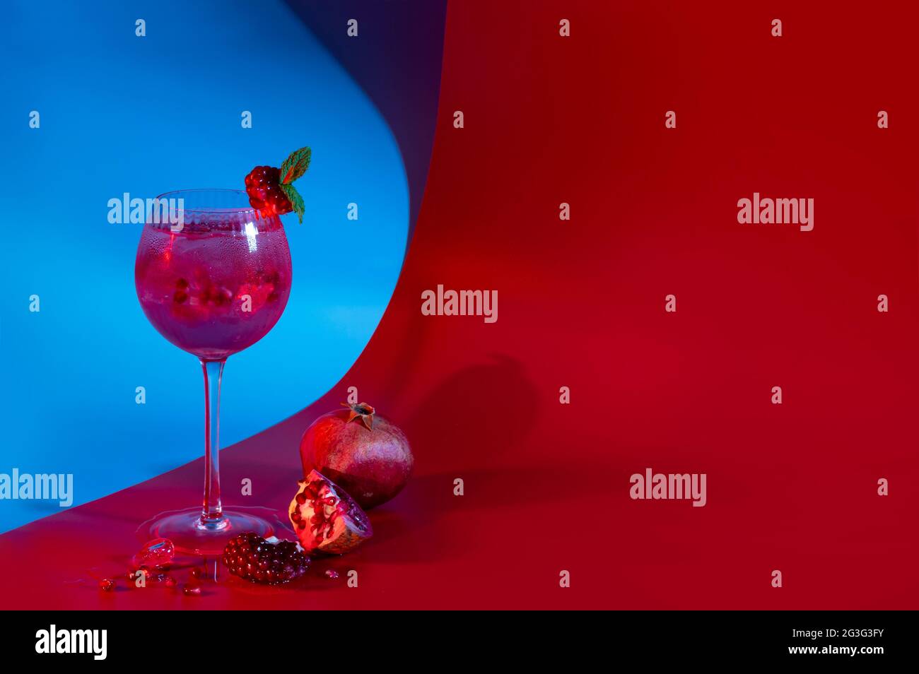 Cocktail di melograno gin e tonico con cubetti di ghiaccio e foglie di menta su sfondo astratto rosso e blu. Spazio di copia neutro Foto Stock