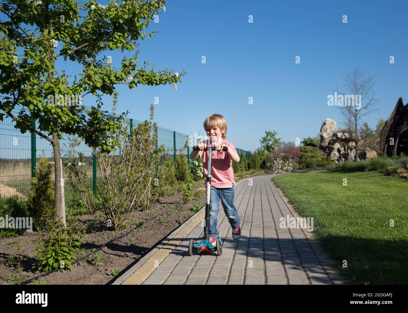 un ragazzo allegro, di 4-5 anni, corre sul marciapiede nel parco su uno scooter. movimento, velocità, attività sportive per bambini Foto Stock