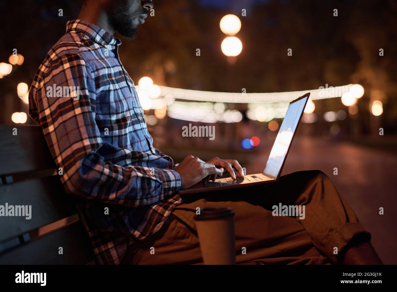 Giovane seduto sulla panchina e lavorando online su un computer portatile nel parco la sera Foto Stock