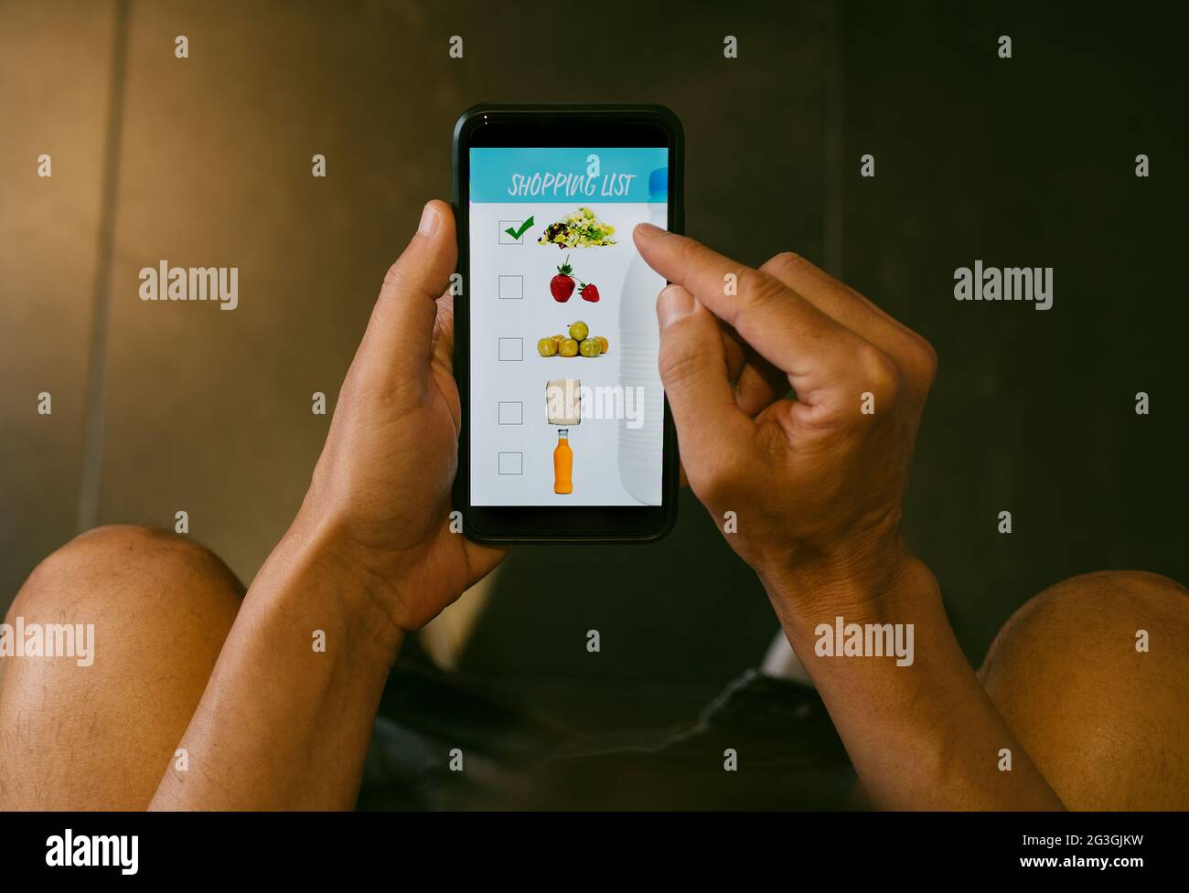 primo piano di un giovane caucasico, seduto nel gabinetto, facendo la sua shopping list nel suo smartphone, con una mock app con immagini di diversi prodotti Foto Stock