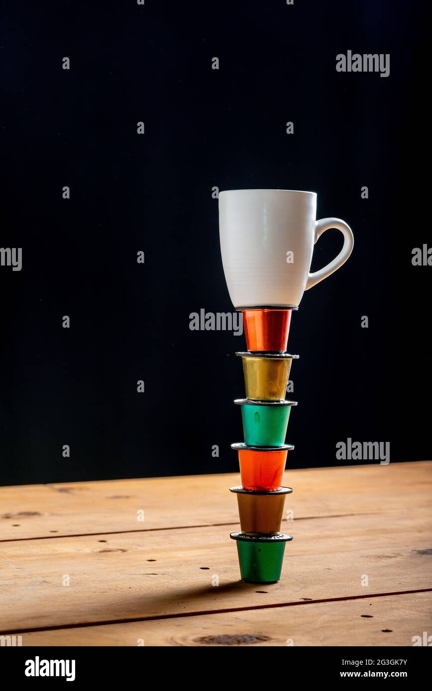 Tazza di caffè in cima a una torre fatta con cialde di caffè colorate e utilizzata su un tavolo di legno su sfondo nero. Foto Stock