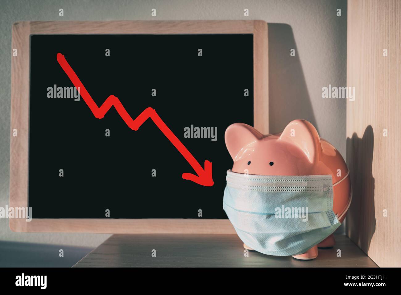 Impatto economico globale della pandemia del coronavirus. Piggy banca indossare maschera chirurgica viso con freccia rossa che mostra crash di mercato su lavagna. Finanziario Foto Stock