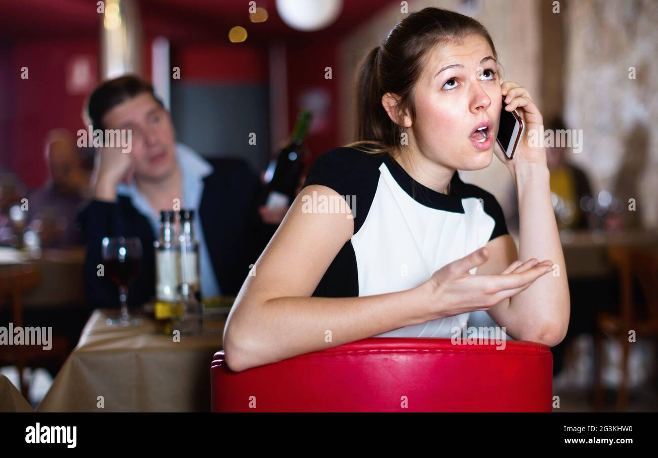 Offesa giovane donna che parla per telefono sullo sfondo con l'uomo ubriaco al tavolo del ristorante Foto Stock