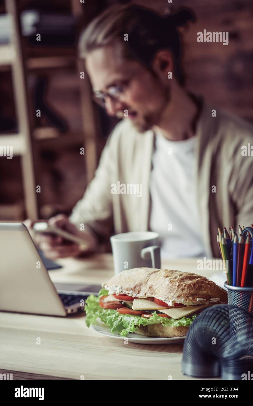 Uomo freelancer a casa, seduto, lavorando andando a mangiare sandwich. Foto Stock