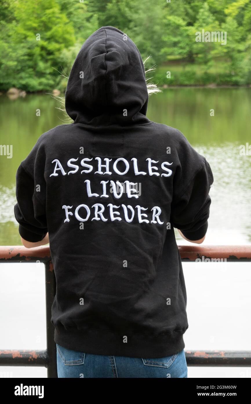Una giovane donna che indossa una felpa con cappuccio che dice che 'Assholes Live Forever'. In un parco a Queens, New York City. Foto Stock