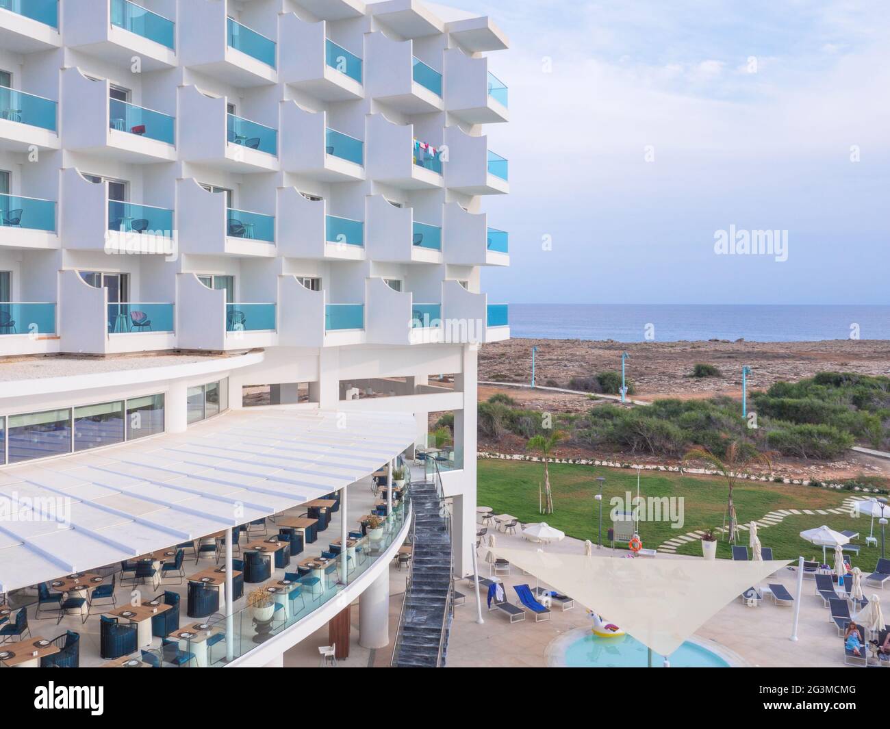 Hotel bianco con file di balconi di plastica blu trasparente e ristorante con terrazza all'aperto con tavoli da pranzo e sedie in legno in attesa di turisti. Foto Stock