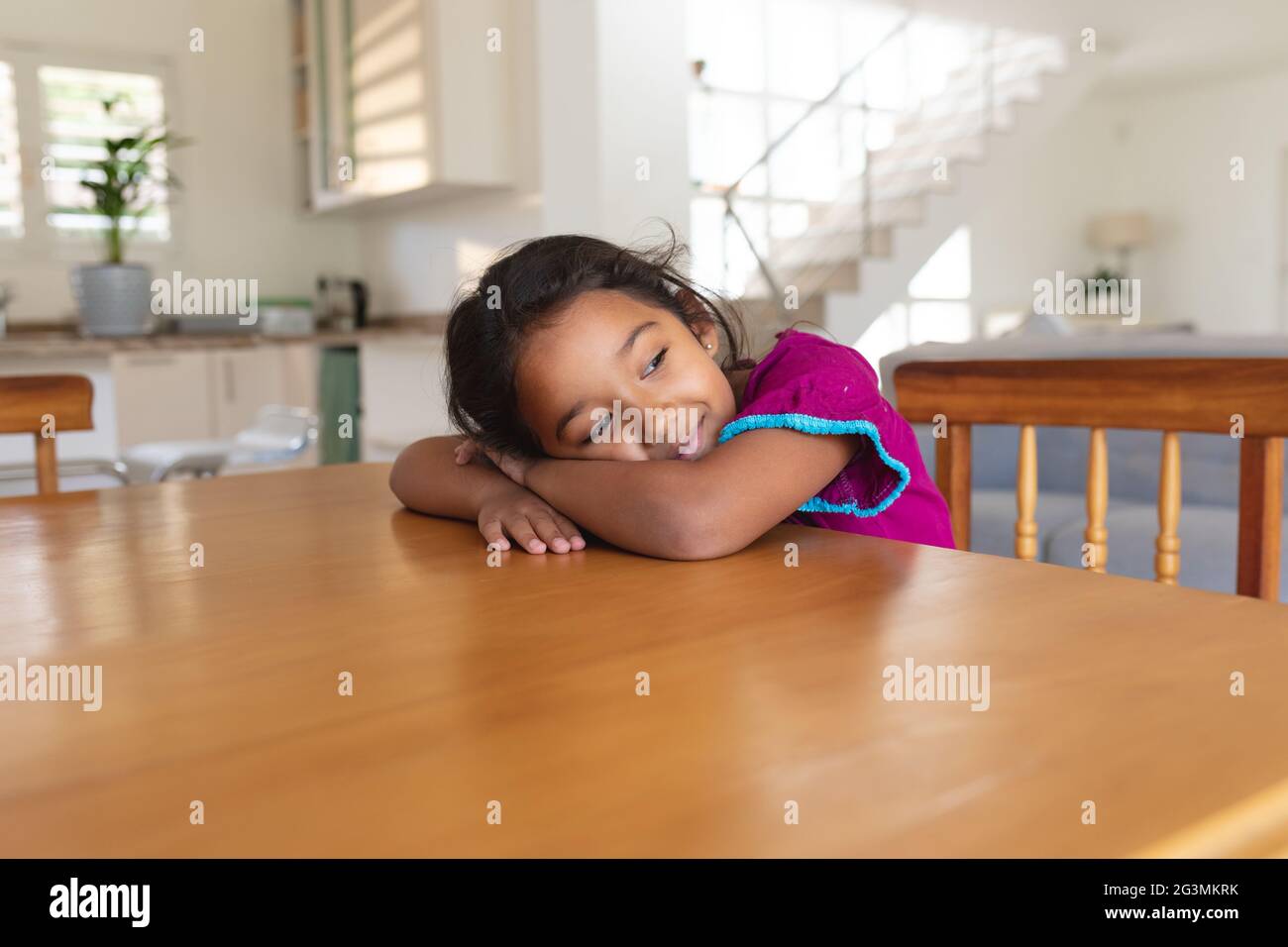 Sorridente ragazza ispanica seduta al tavolo in cucina appoggiata alla testa di ams, guardandosi via Foto Stock