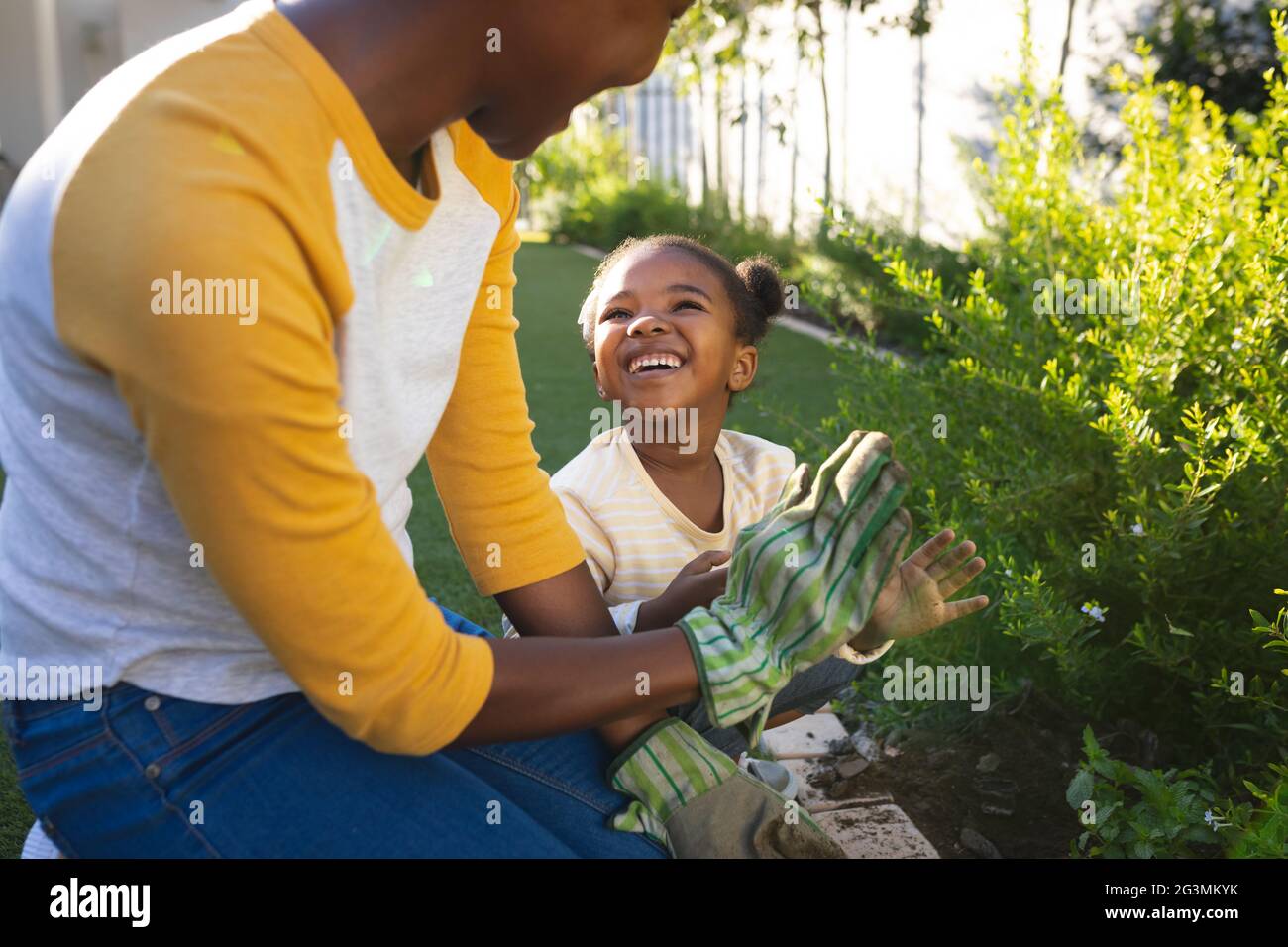 Sorridente madre afroamericana e figlia inginocchiati tendendo a piante in giardino soleggiato Foto Stock