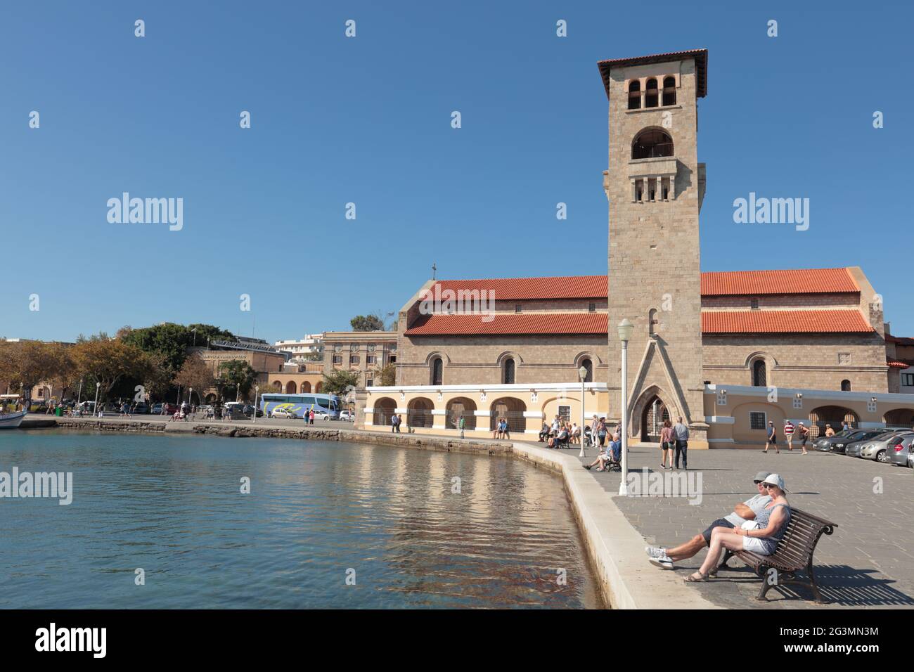 Persone che riposano al porto di Mandraki di Rodi, isola di Rodi, Grecia contro la Chiesa di Annunciazione della Vergine nella città di Rodi, Dodecanese Foto Stock
