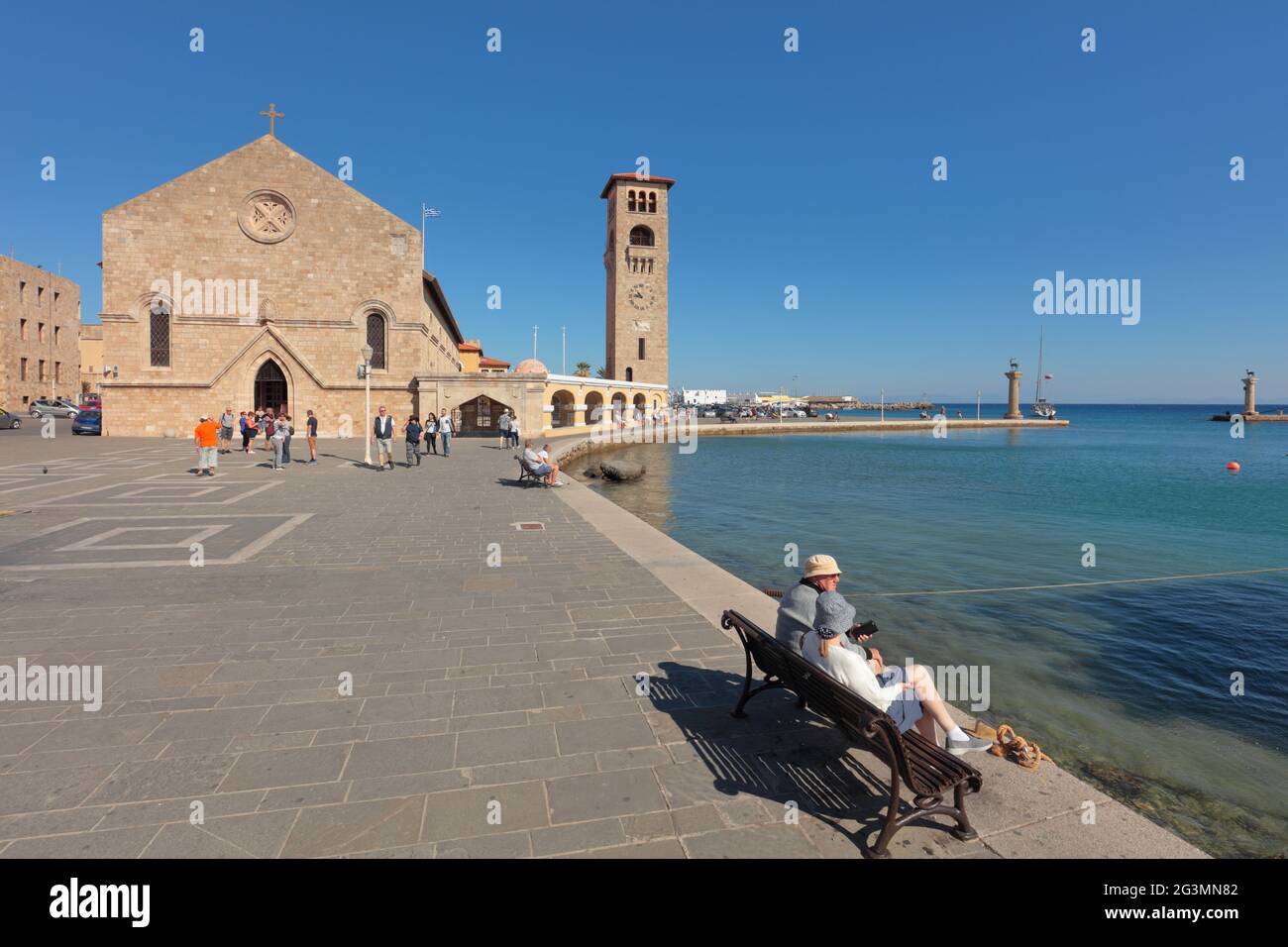 Persone che riposano al porto di Mandraki di Rodi, isola di Rodi, Grecia contro la Chiesa di Annunciazione della Vergine nella città di Rodi, Dodecanese Foto Stock
