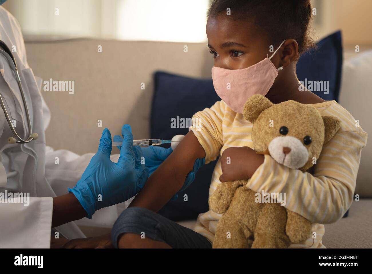 African american femmina medico che dà la vaccinazione di covid alla paziente della ragazza in maschera di faccia a casa Foto Stock