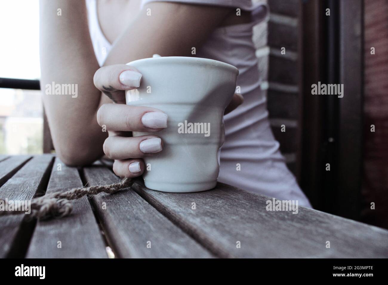 La mano della donna sta tenendo una tazza di caffè di porcellana dissagomata, poggiando il gomito su un tavolo di legno bella manicure Foto Stock