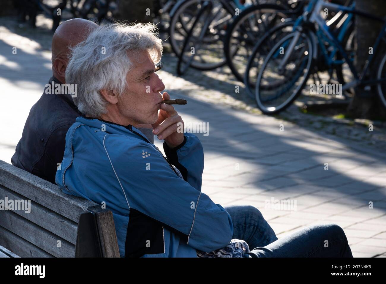 L'uomo si siede su una panchina di legno con un altro uomo e fuma un sigaro. Vista laterale retroilluminata, una fila di biciclette sullo sfondo Foto Stock