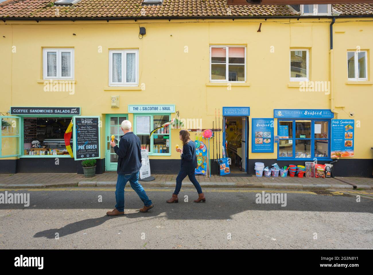 Negozi di Salcombe, vista in estate di una coppia mangiare gelati, come si passa una fila di negozi colorati nel centro di Salcombe, Devon, Inghilterra, Regno Unito Foto Stock