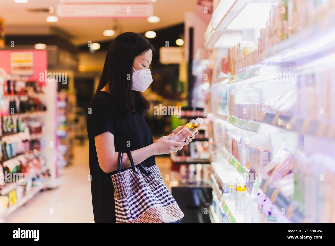 Donna asiatica in maschera protettiva che tiene una bottiglia di shampoo nel supermercato. Foto Stock