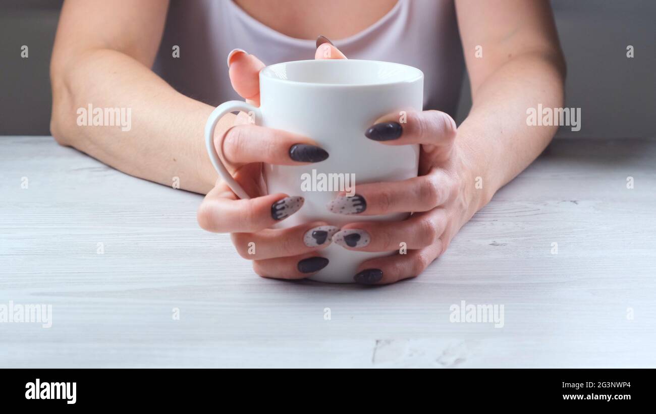 Le mani femminili stanno tenendo una tazza bianca di caffè su un tavolo di legno chiaro. Il concetto di sofferenza, disperazione, problemi Foto Stock
