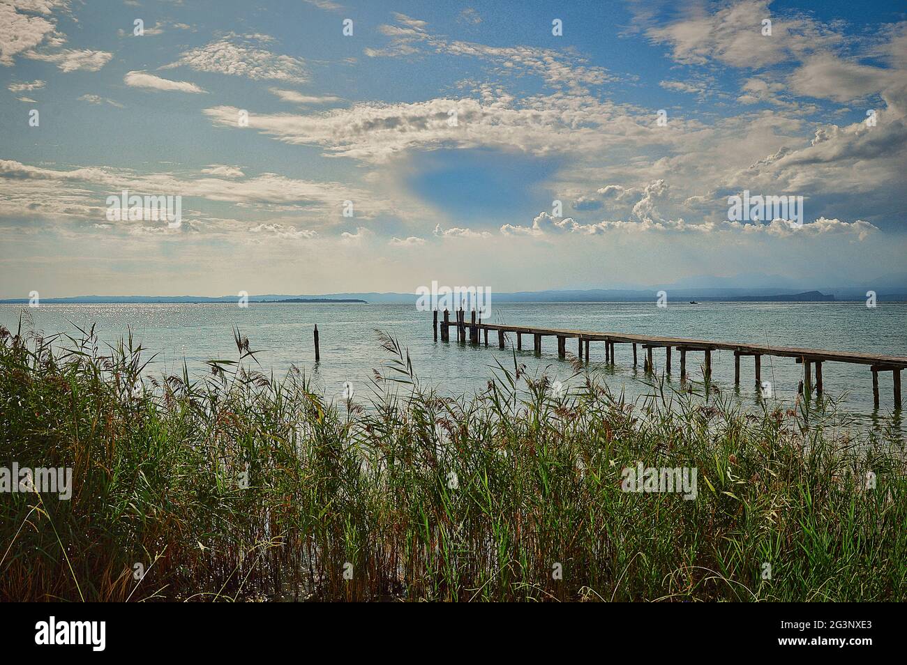 Spiaggia naturale/molo/spiaggia del lago di Garda con ponte, erba, acqua e cielo blu drammatico Foto Stock