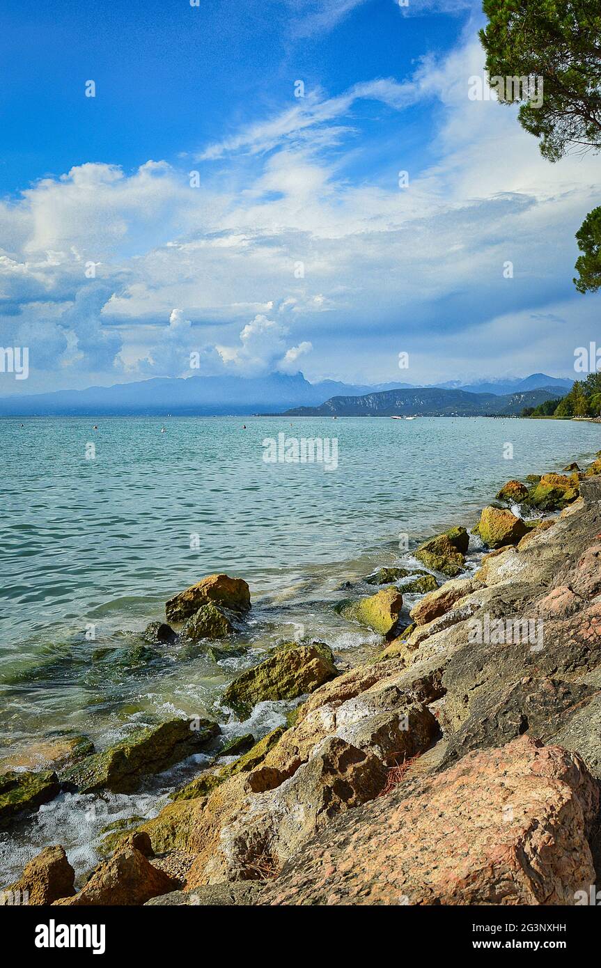 Paesaggio di riva / molo / spiaggia del lago di Garda con acqua blu, rocce, e cielo blu drammatico Foto Stock
