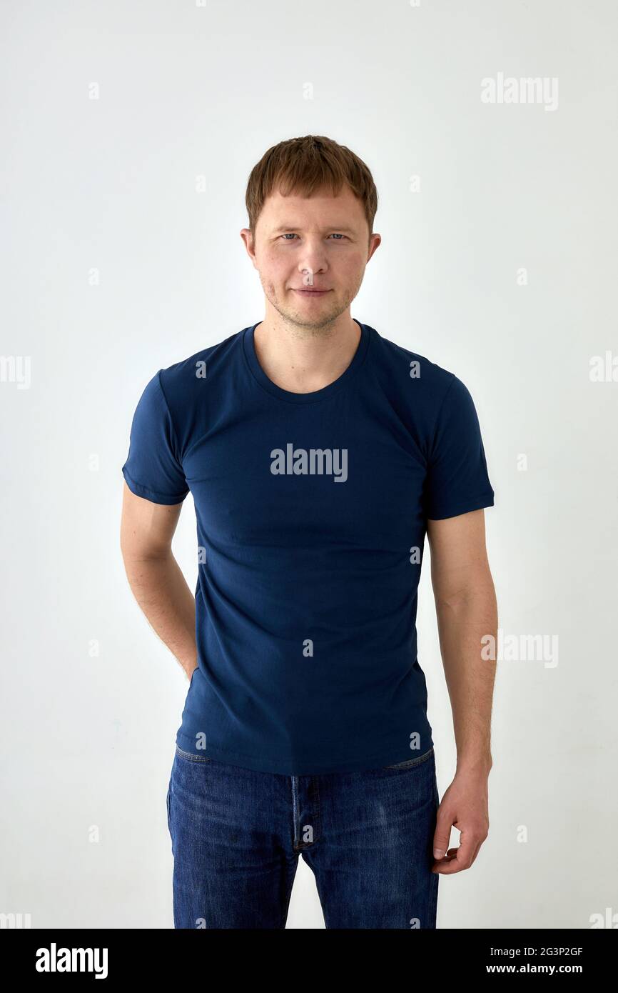 Giovane uomo positivo in jeans con la mano dietro e la t-shirt blu scuro in piedi su sfondo bianco e guardando la macchina fotografica Foto Stock