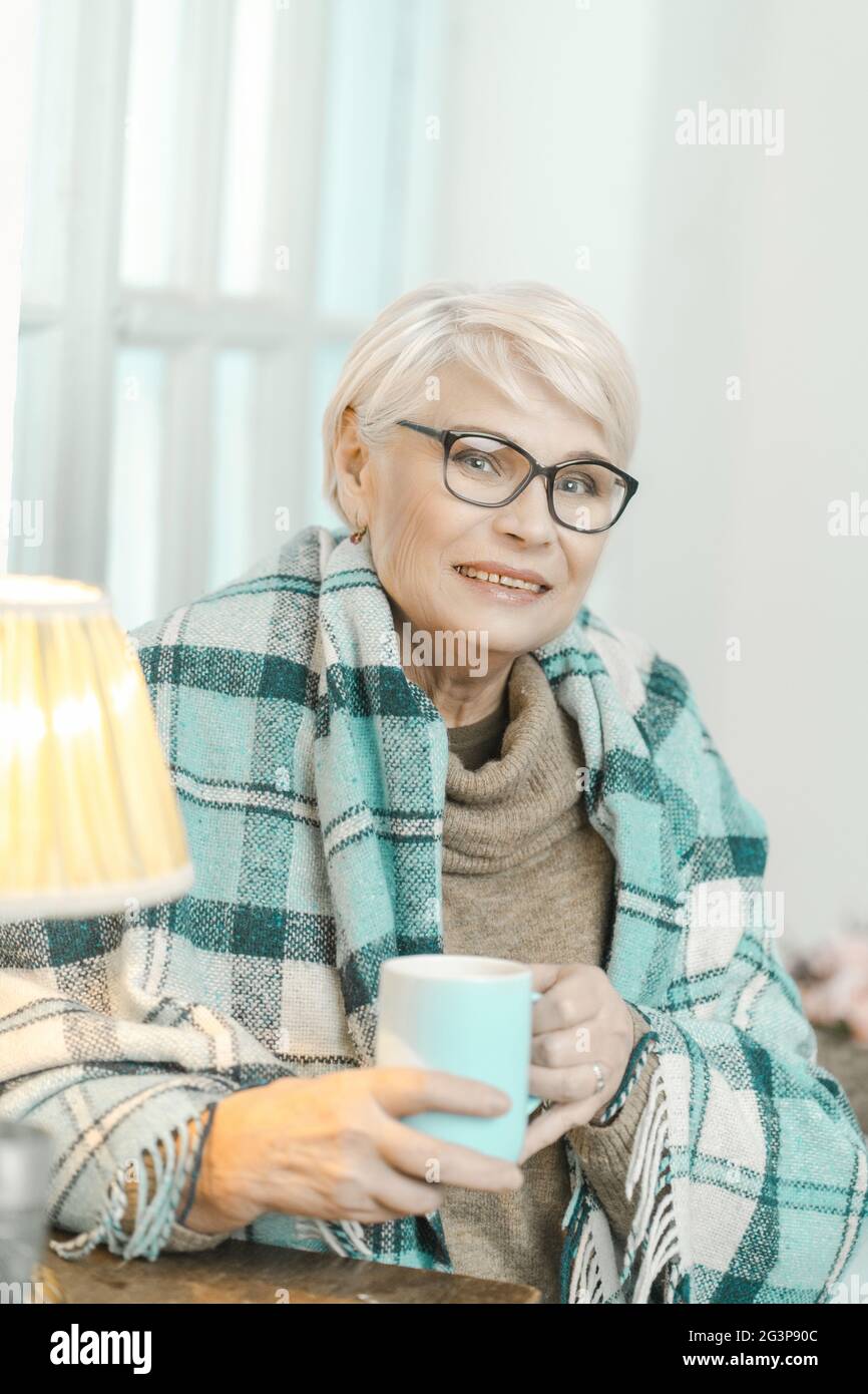 Donna anziana avvolta in una quadri a scacchi sta bevendo il tè a casa Foto Stock
