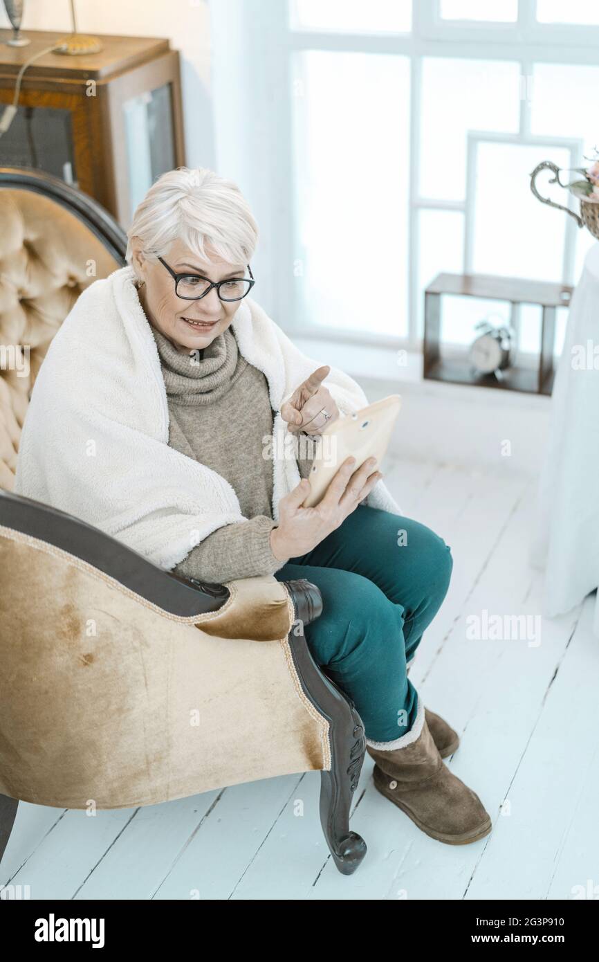 Vecchia donna dai capelli grigi che riposa a casa con una tavoletta in mano. Foto Stock