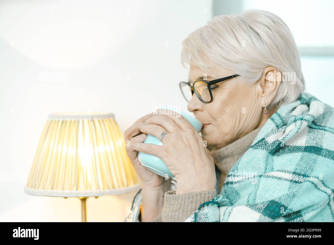 Donna anziana avvolta in una quadri a scacchi sta bevendo il tè a casa Foto Stock