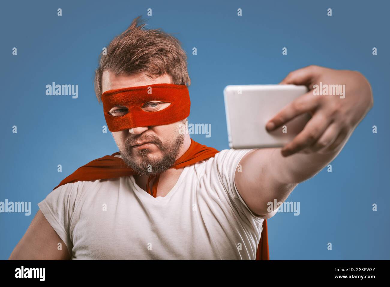 Super Hero uomo prende selfie da telefoni cellulari. Uomo sicuro in un costume super eroe in una maschera rossa e mantello posa su jeans sbiaditi Foto Stock