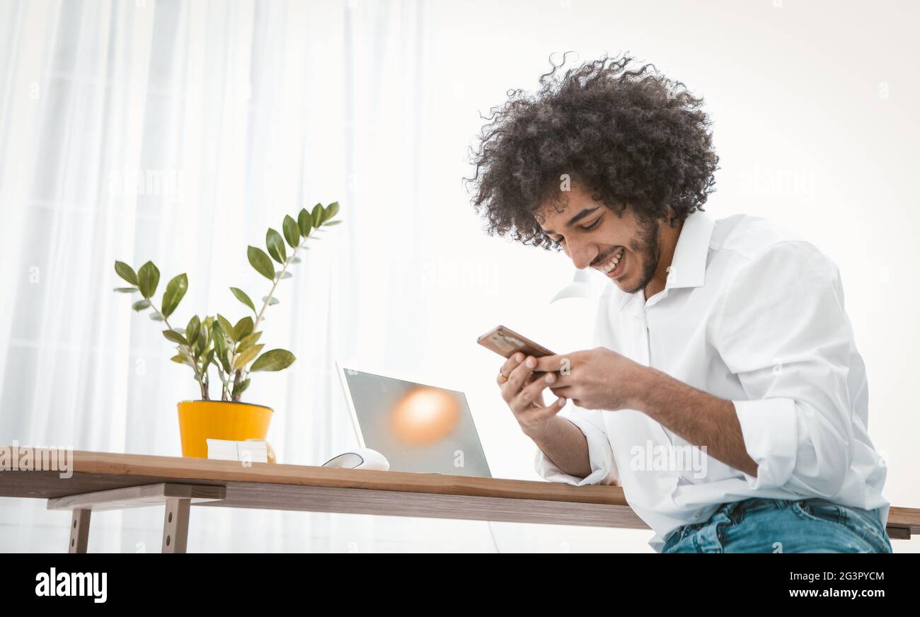 Uomo d'affari sorridente che parla sul telefono cellulare con amici, partner d'affari o fidanzata, seduto su una scrivania di legno. Vista laterale Foto Stock