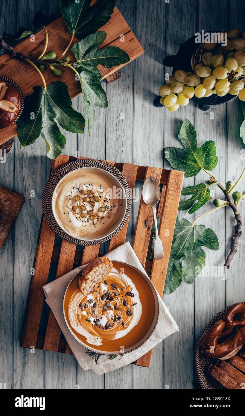 Fotografia piatta di una zuppa di crema vegana su un tavolo di legno vecchio. Vista dall'alto di una purea di prodotti di stagione su un vassoio di legno tra foglie di fico, pane A. Foto Stock