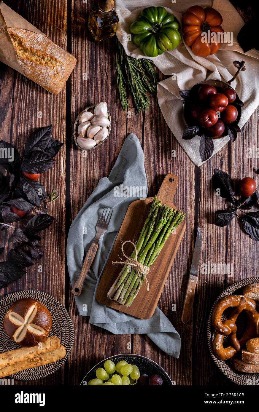 La foto scura di asparagi, pane, pomodori, aglio e prugne è piatta su un tavolo di legno antico. Ricetta di verdure per autunno o inverno. Moody top Foto Stock