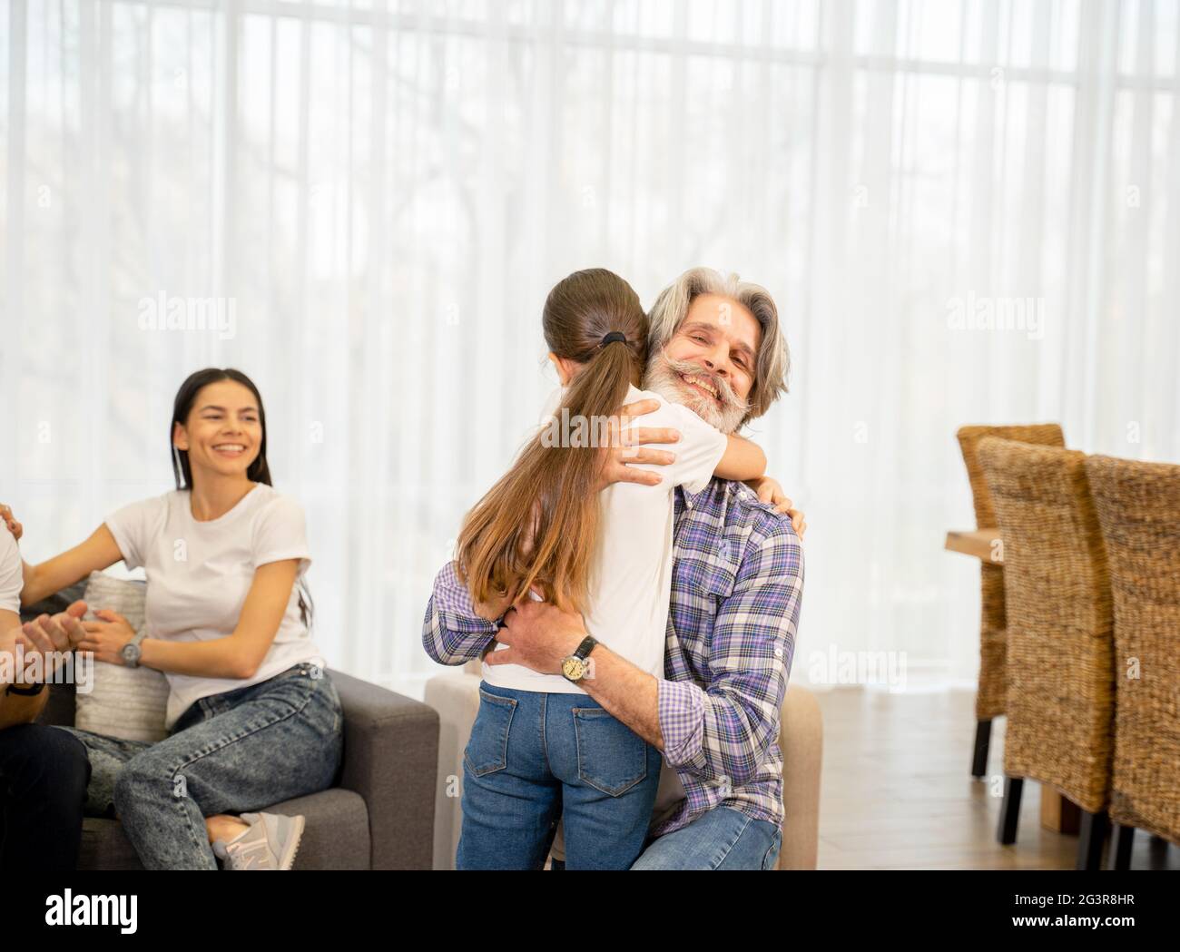 Eccitata nipotina carina hugging nonno felice che esprime l'amore quando si passa il tempo insieme e giocare in salotto leggero, madre felice che li guarda e sorridendo. Concetto di famiglia Foto Stock