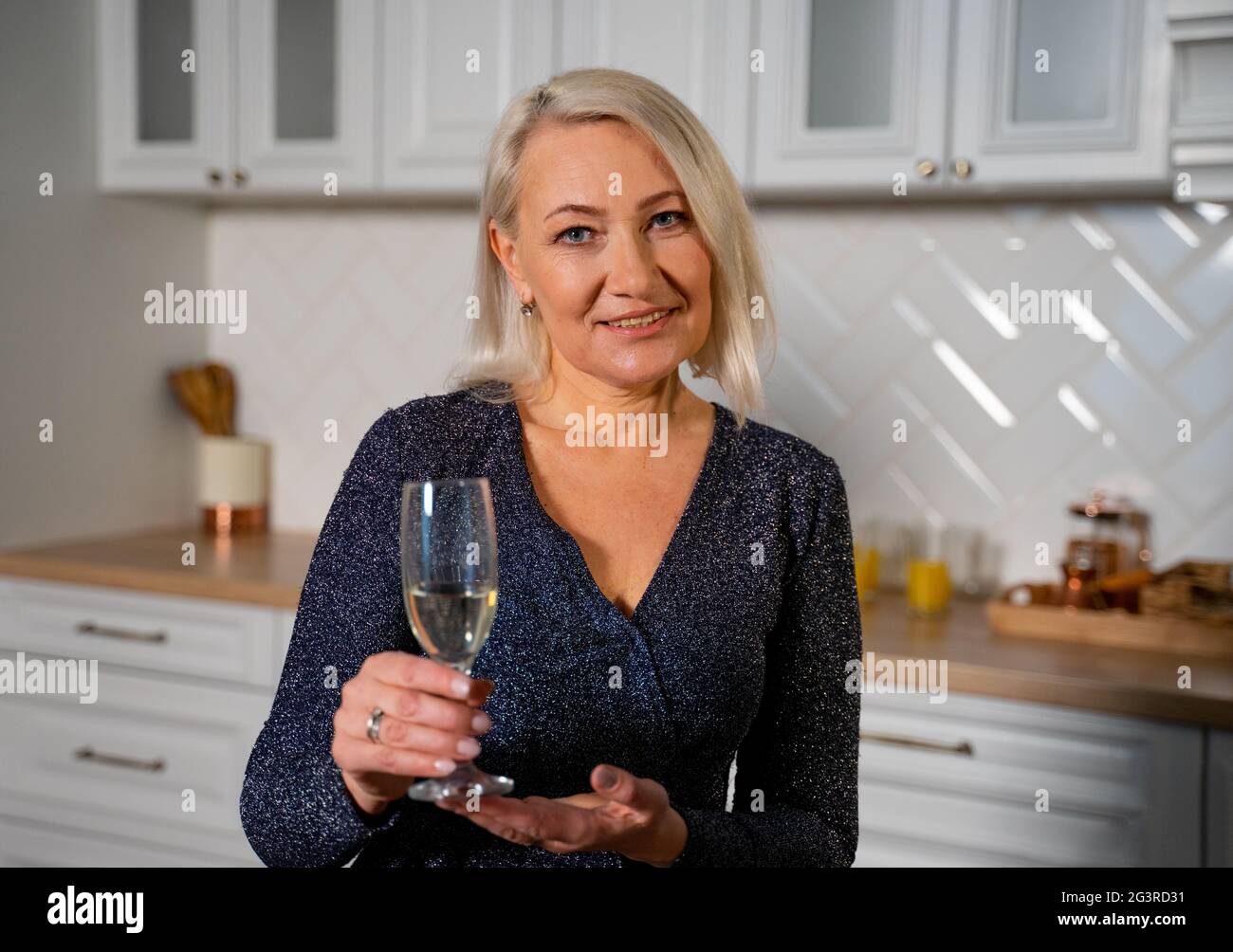 Ritratto di nonna sorridente in bel vestito festivo in piedi in elegante  cucina che tiene un bicchiere di champagne che progetta di fare un brindisi  e Wish Buon compleanno. Concetto di celebrazione