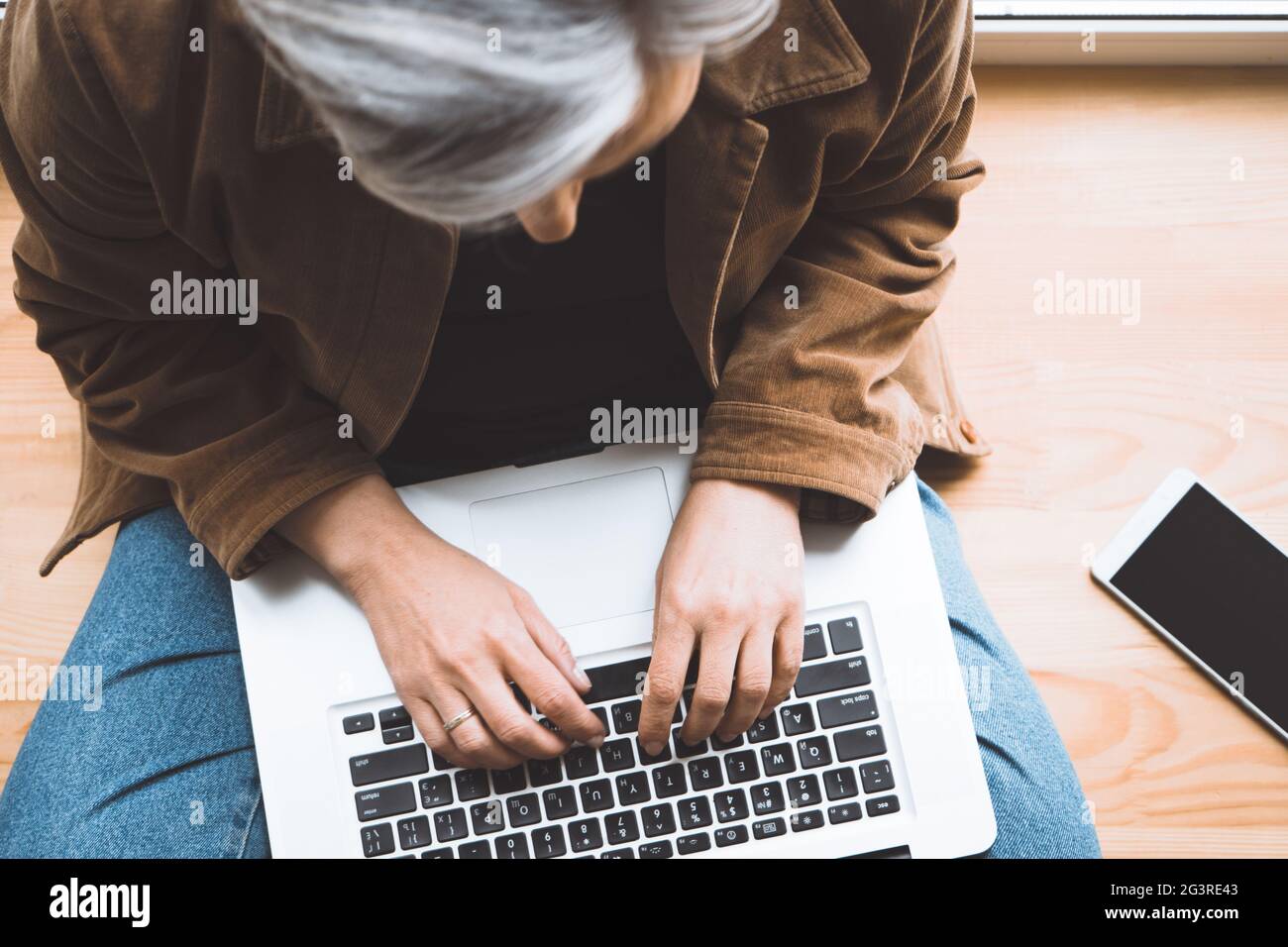Donna grigia che dattilografa la tastiera del laptop seduta su windowsill di legno con il telefono cellulare su di esso. Vista dall'alto. Focus selettivo su fema Foto Stock