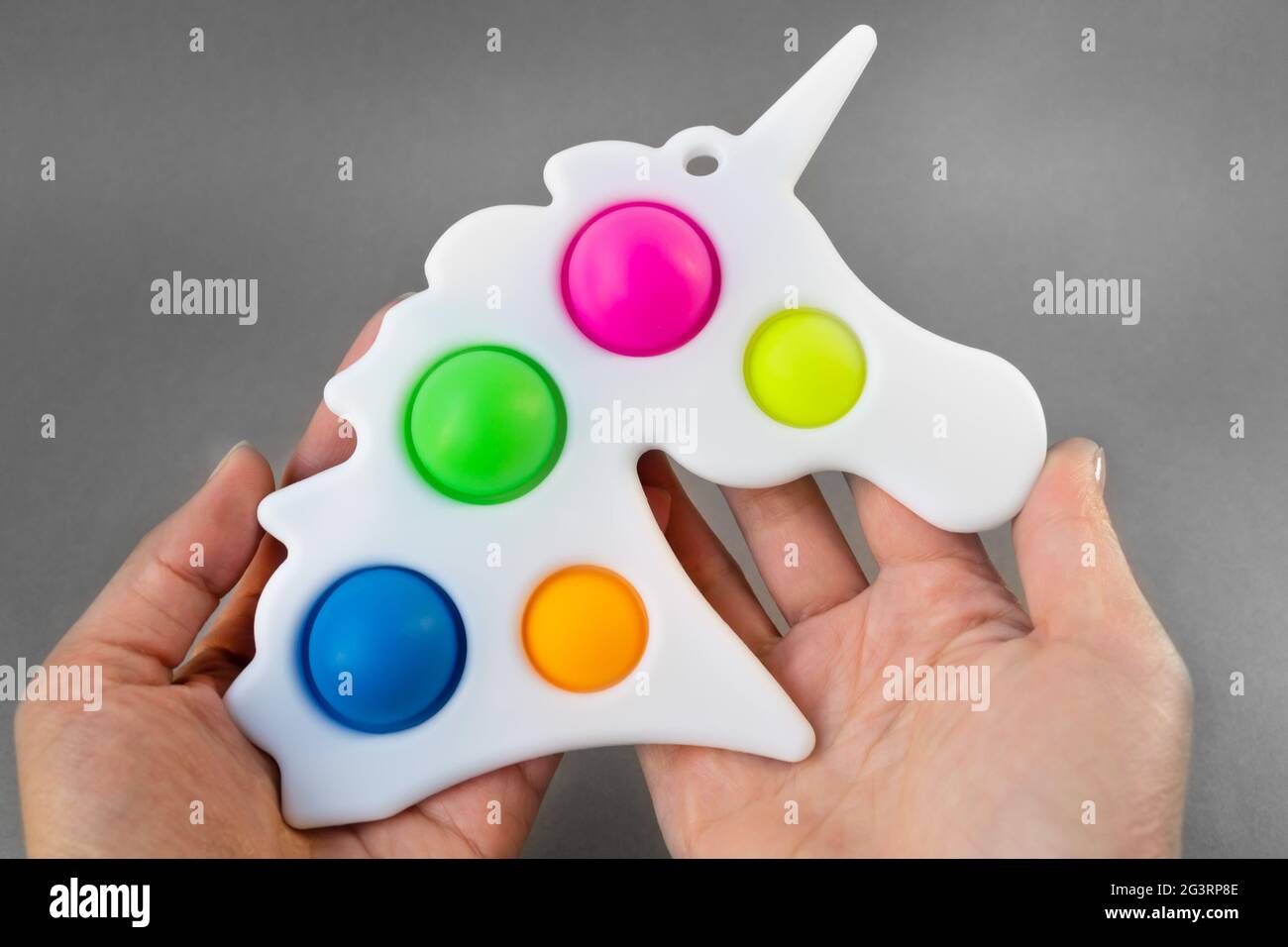 Giocattolo luminoso e colorato semplice unicorno Dimple. Popolare giocattolo antistress su sfondo grigio nelle mani di una donna Foto Stock