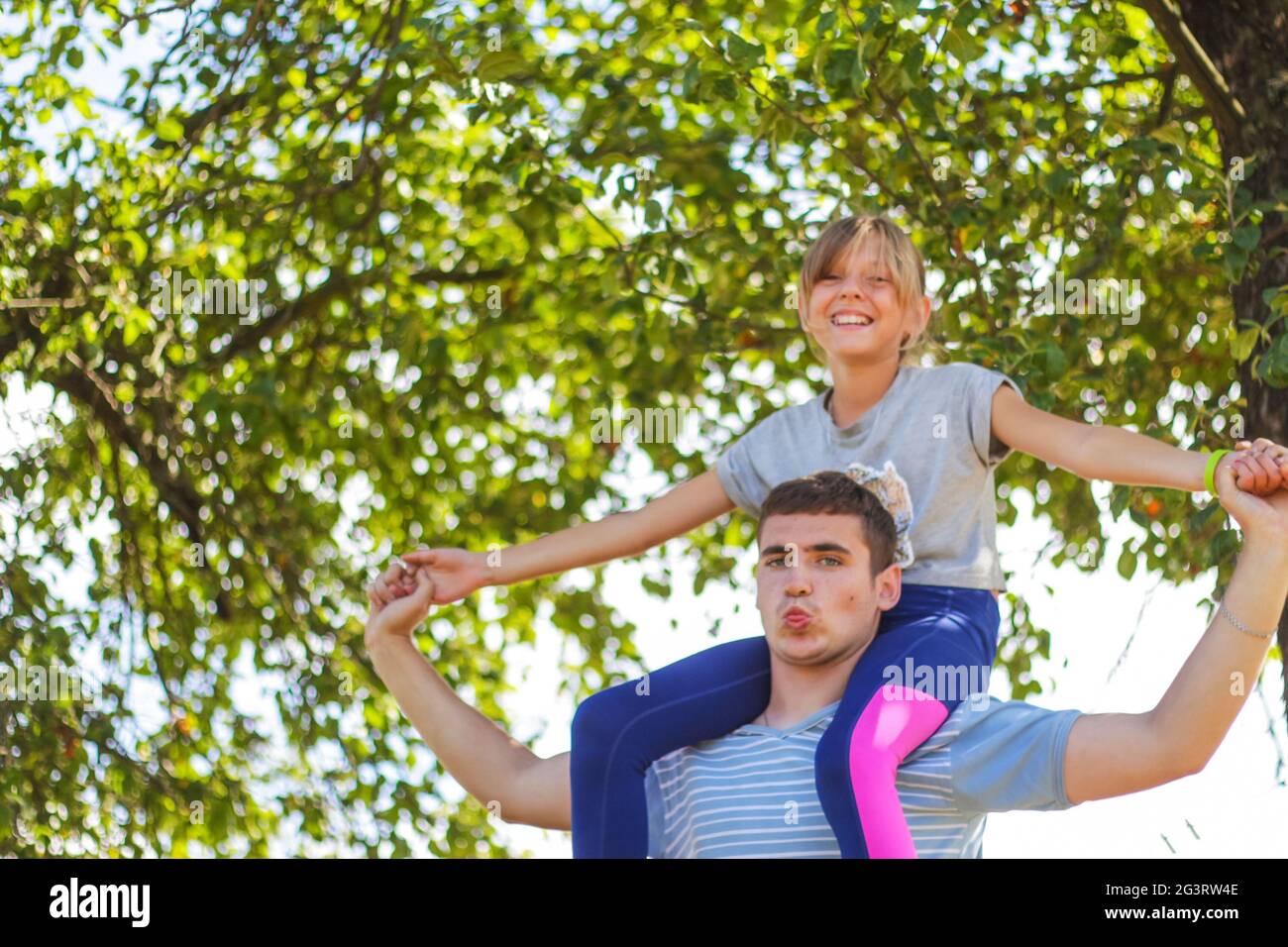 Fratello disinnescato che cavalcava la sorella sulla schiena. Ritratto di ragazza felice sulle spalle dell'uomo, piggyback. La ragazza vola, alza la mano. Famiglia che gioca all'esterno. Albero verde Foto Stock