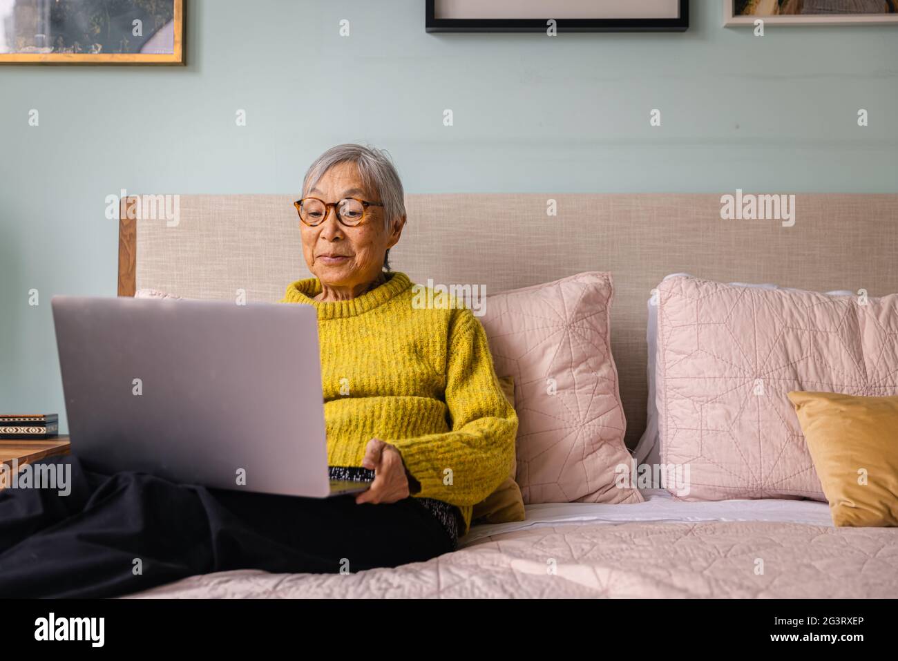 Donna asiatica anziana che usa il computer portatile mentre si siede in camera da letto a casa Foto Stock