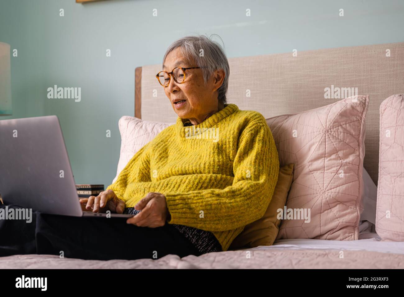Donna asiatica anziana che usa il computer portatile mentre si siede in camera da letto a casa Foto Stock