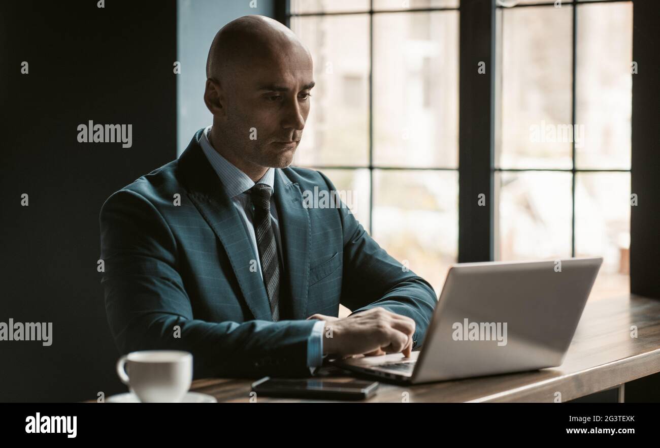 Uomo d'affari baldone di mezza età in business suite che lavora su un computer portatile accanto a finestre aperte in un ufficio moderno. Bel lavoro uomo audace Foto Stock