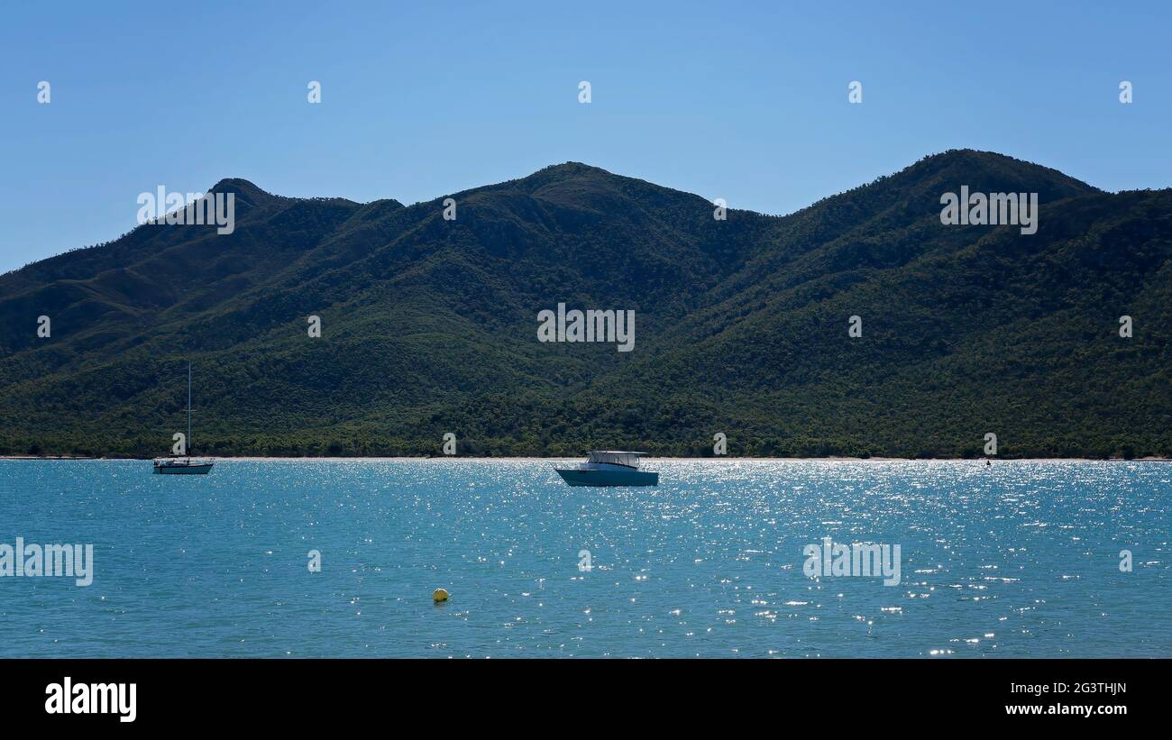 una barca a motore e uno yacht ormeggiati al largo della costa su un mare calmo che brildisca la luce del mattino presto su uno sfondo di montagna Foto Stock
