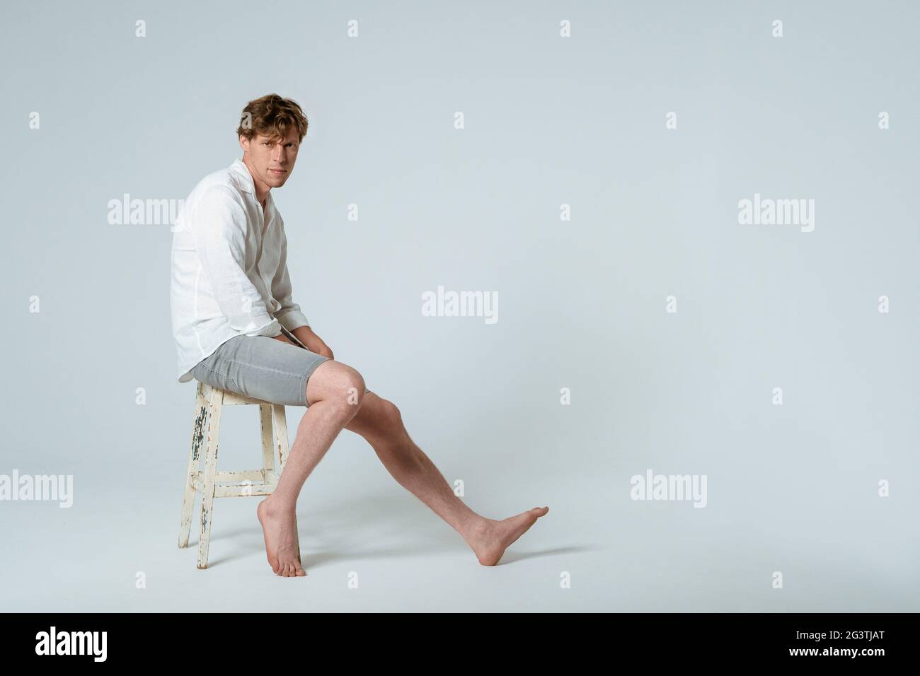 Foto a lunghezza intera di un giovane bel ragazzo seduto su una sedia che indossa una camicia bianca e pantaloncini grigi con una gamba che guarda circa Foto Stock