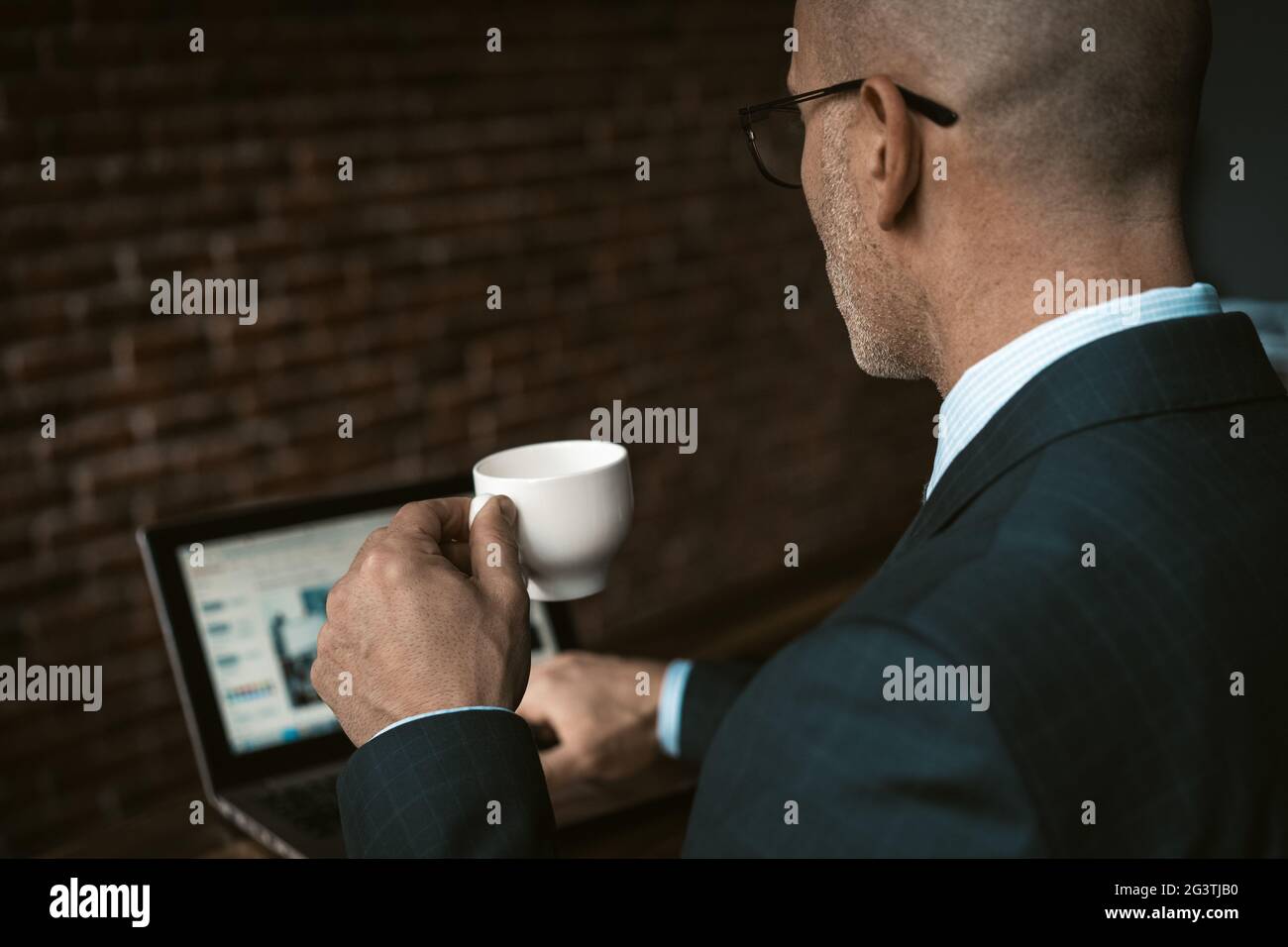 Vista posteriore di un uomo d'affari maturo in un tuta da lavoro, tenendo una tazza di caffè seduto al tavolo mentre si lavora su un computer portatile e sul retro Foto Stock