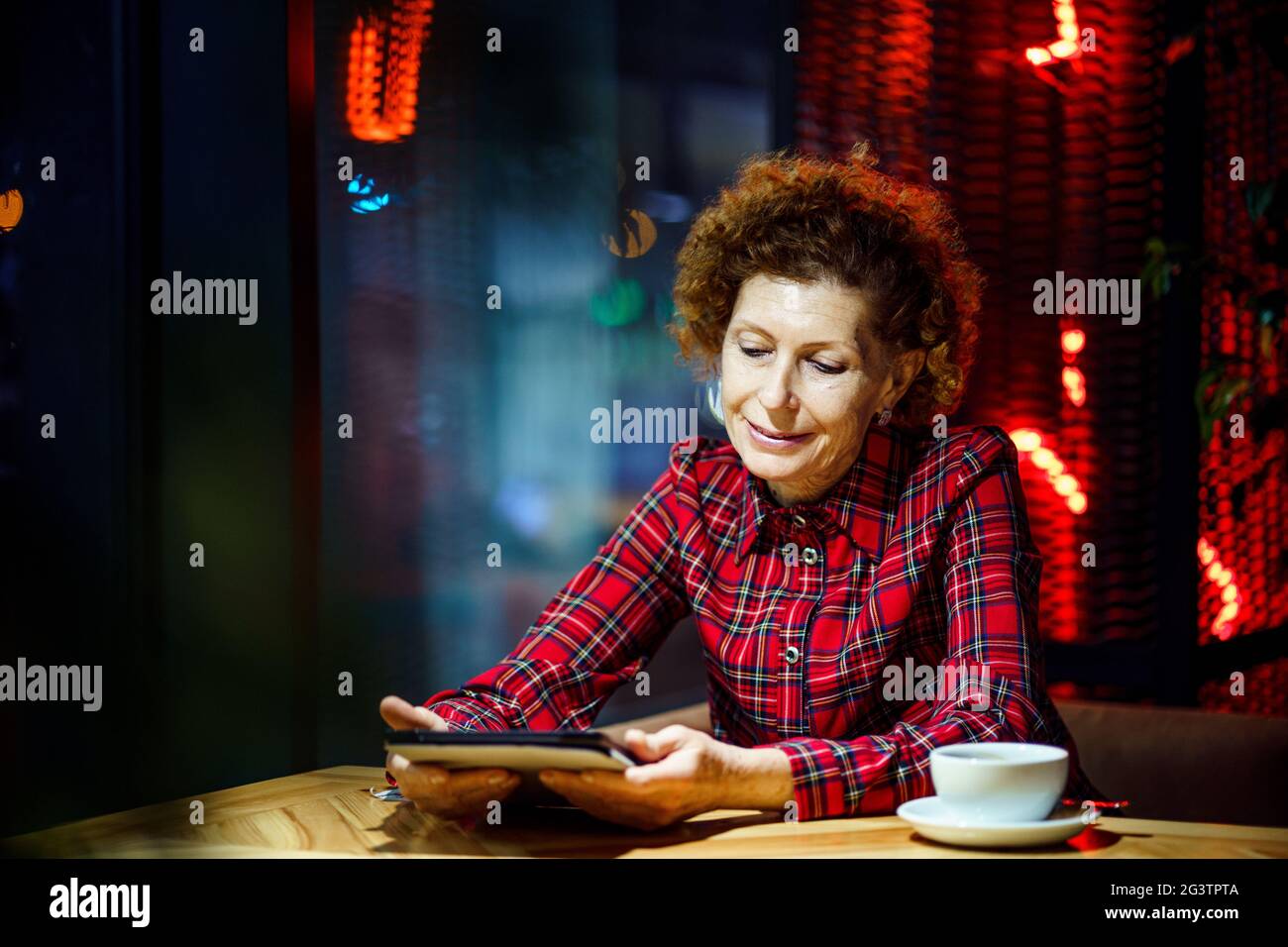 Donna matura con capelli ricci biondi controlla la posta e le notifiche dei social media tramite tablet, sorridente donna che tiene il tablet, watchin Foto Stock