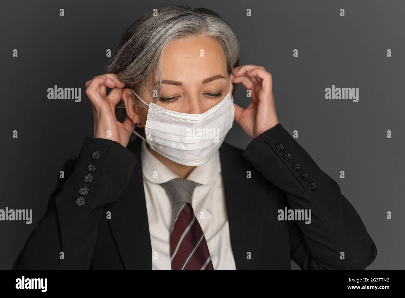 Donna d'affari di mezza età con capelli grigi che si appone su una maschera medica protettiva bianca che guarda verso il basso. Ritratto della donna anziana moderna Foto Stock