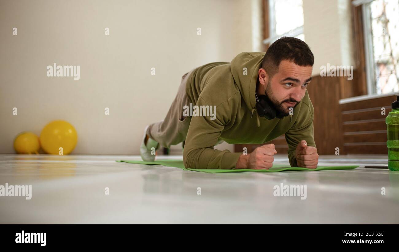 Giovane uomo sportivo che fa esercizio a gomito in palestra vuota con palline da palestra gialle dietro di lui tutto in tuta di colore kaki. Muscola Foto Stock