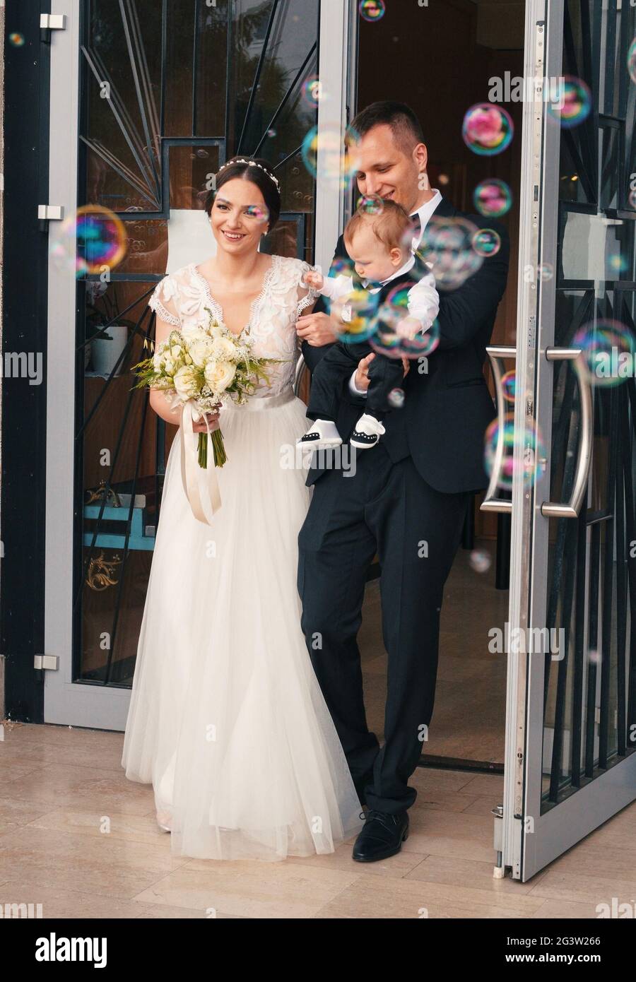 Appena sposato coppia con appena battezzato bambino uscendo dalla chiesa con bolle di sapone di fronte a loro Foto Stock